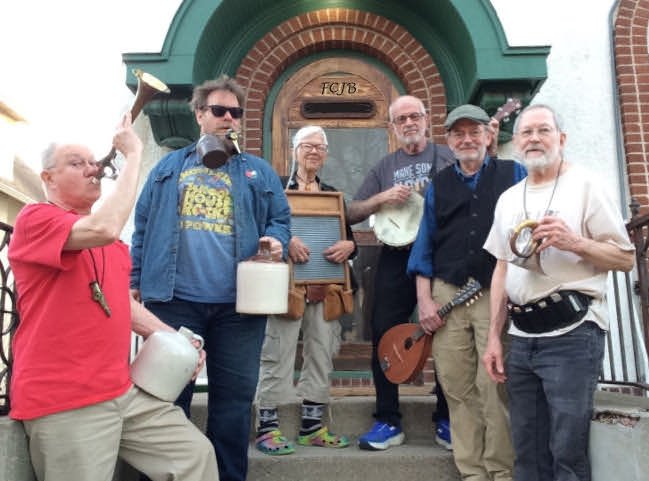 The six members of the Fat Chance Jug Band pose with a jug, washboard, banjo, mandolin and guitars on a brick doorstep.
